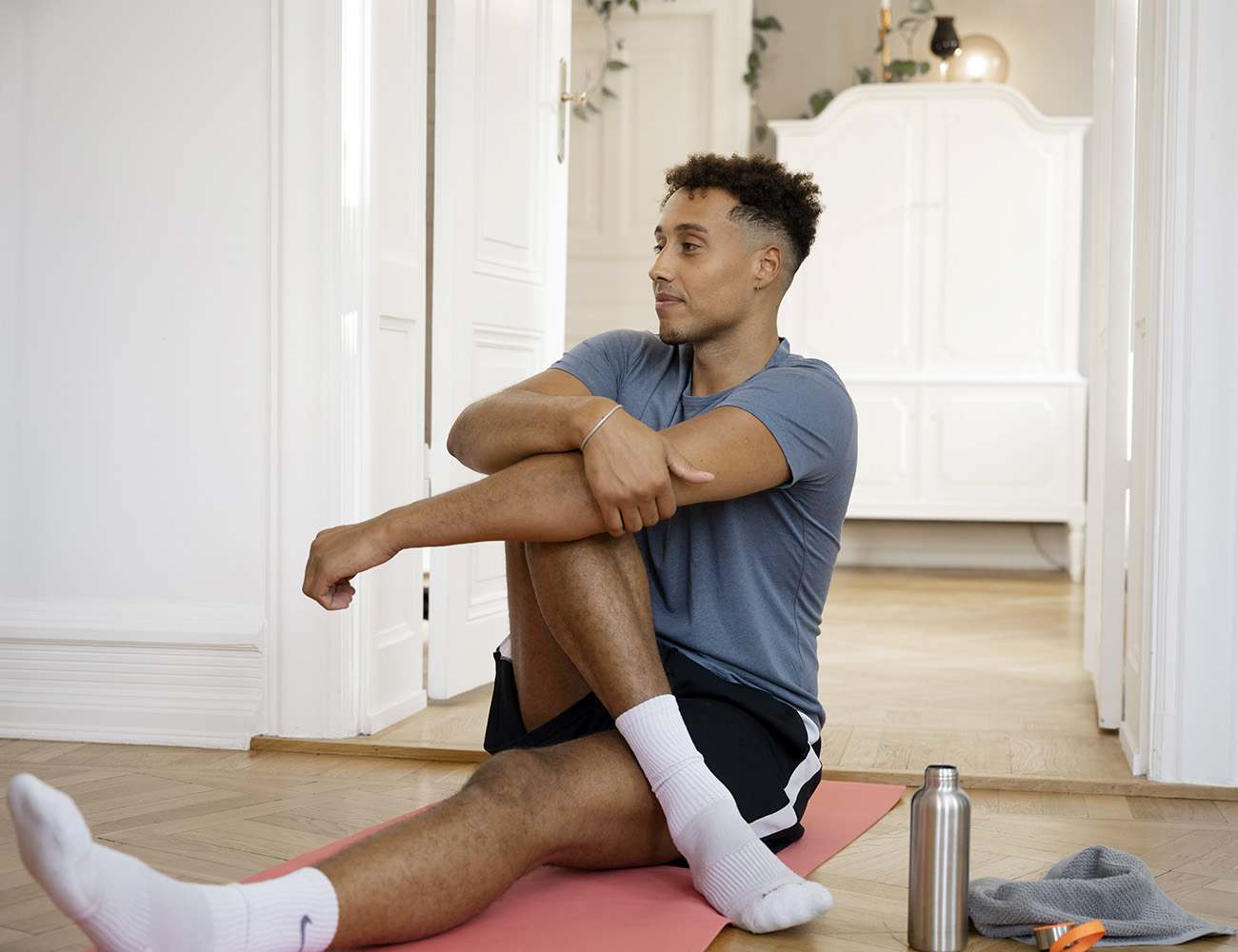 En kille med kort mörkt hår, blå tshirt och ett par svarta shorts. Sitter på en rosa yogamatta på golvet i sitt hem. Bredvid ser man en vattenflaska i plåt och en grå liten handduk.
