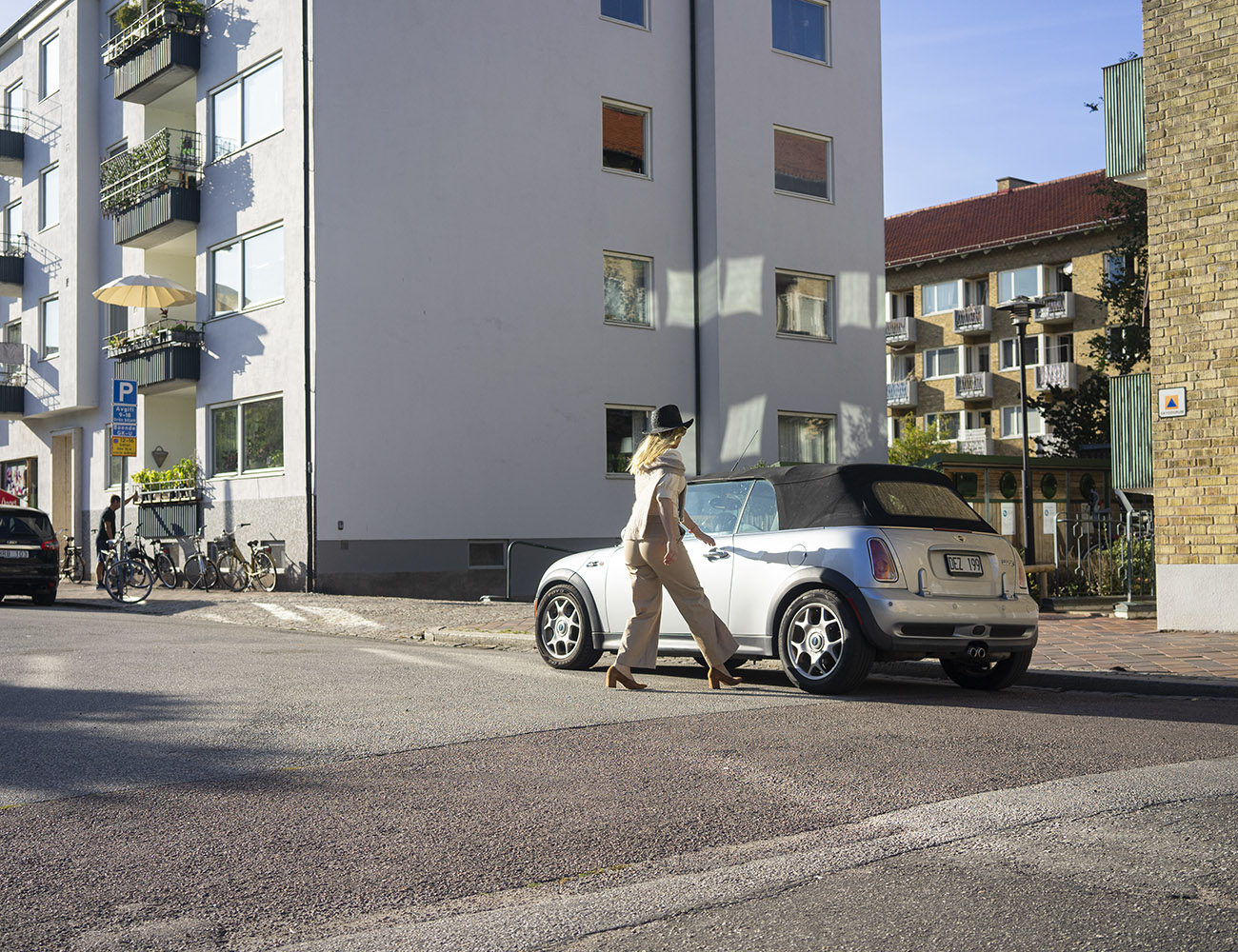 Nainen kävelee kadulle pysäköityyn hopeanväriseen autoonsa. Takana näkyvät valkoinen ja keltainen talo.