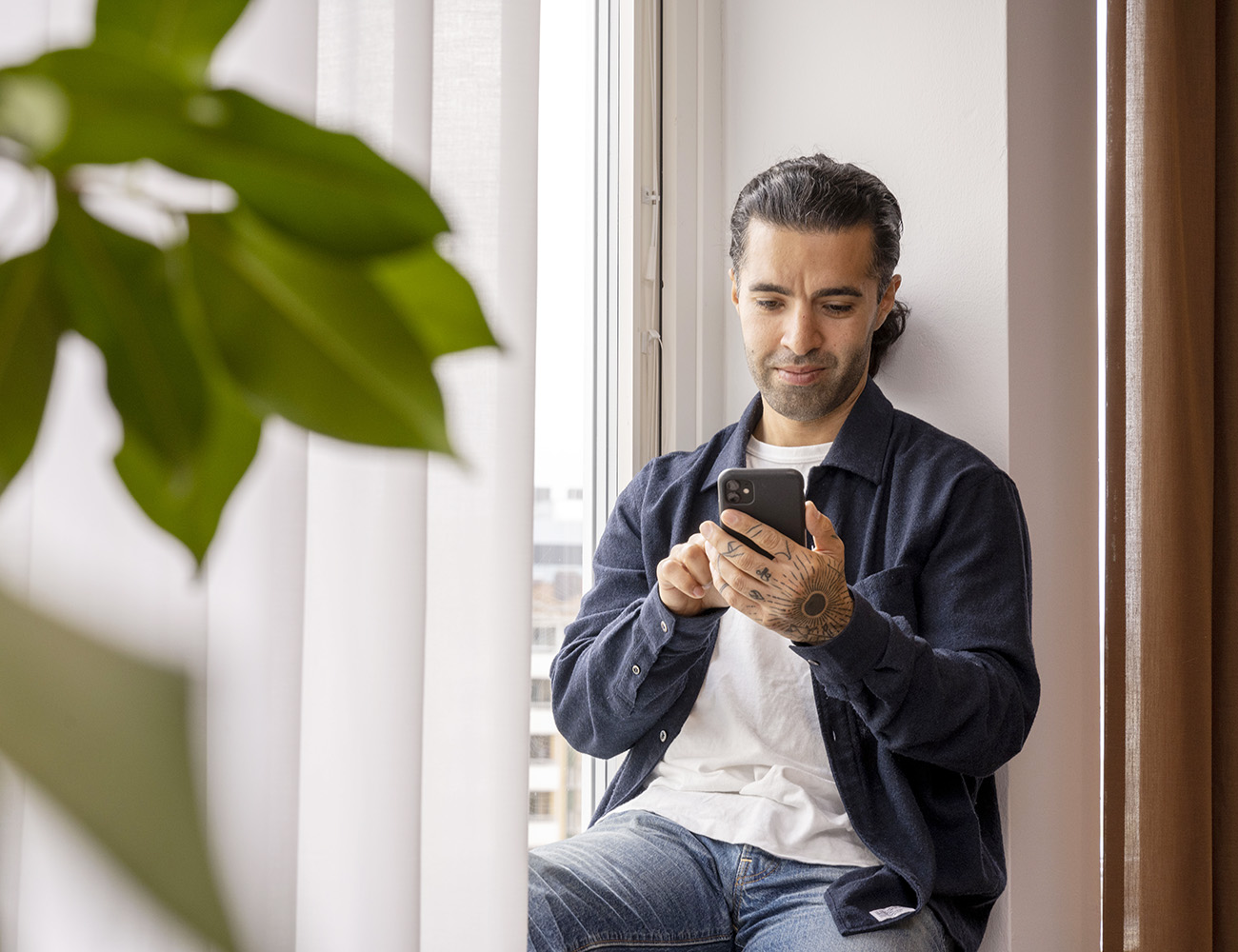 En kille med mörkt hår och skägg sitter i ett fönster och kollar på sin mobil. I fönstret hänger ett par ljusa gardiner och framför står en stor grön växt.