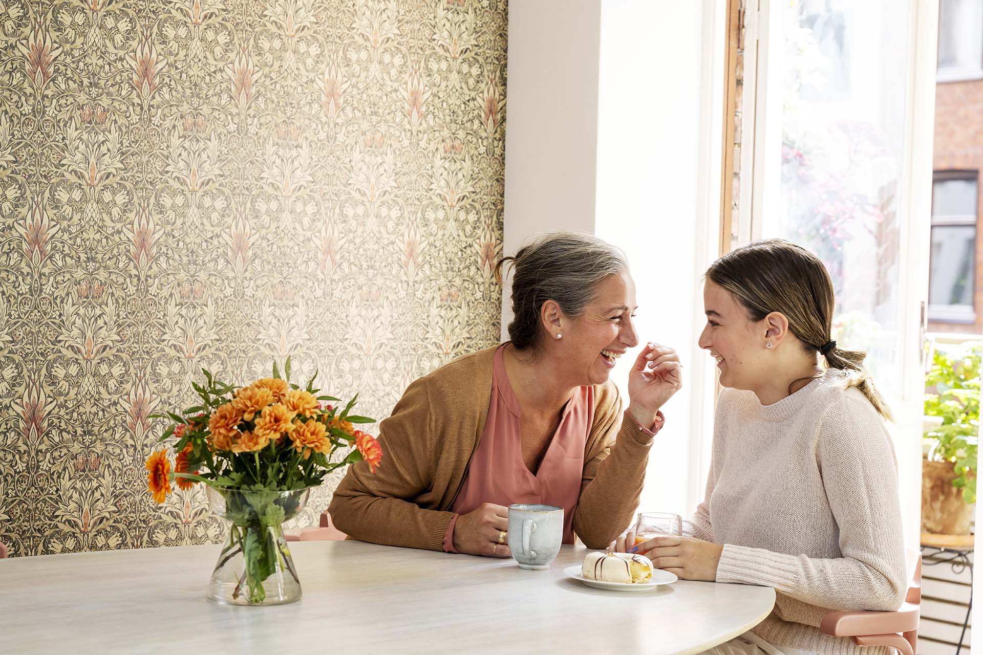 En kvinna i rosa tröja sitter tillsammans med en flicka i vit tröja vid ett köksbord. Bakom ser man en mönstrad tapet och på köksbordet står det en glasvas med orange snittblommor.