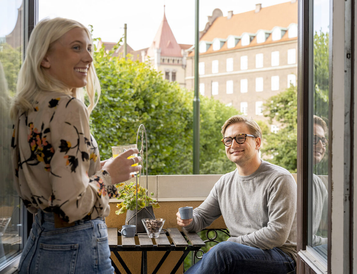 En tjej med ljust hår och en blommig blus står på en balkong. En kille med grå tröja och glasögon sitter på balkongen och dricker en kopp kaffe och tittar på tjejen.