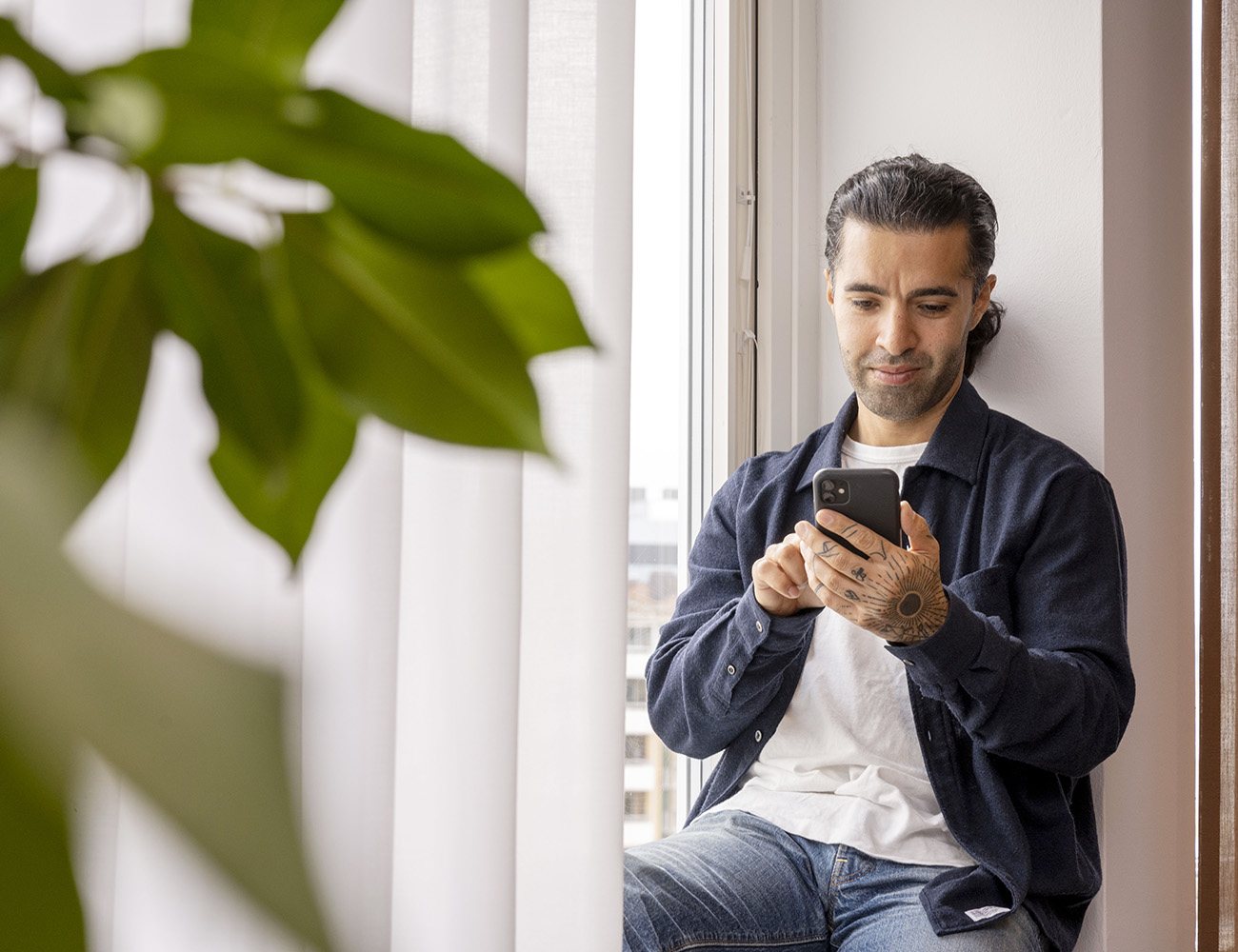 En man med tatueringar på sin hand sitter i fönstret och kollar på sin mobil