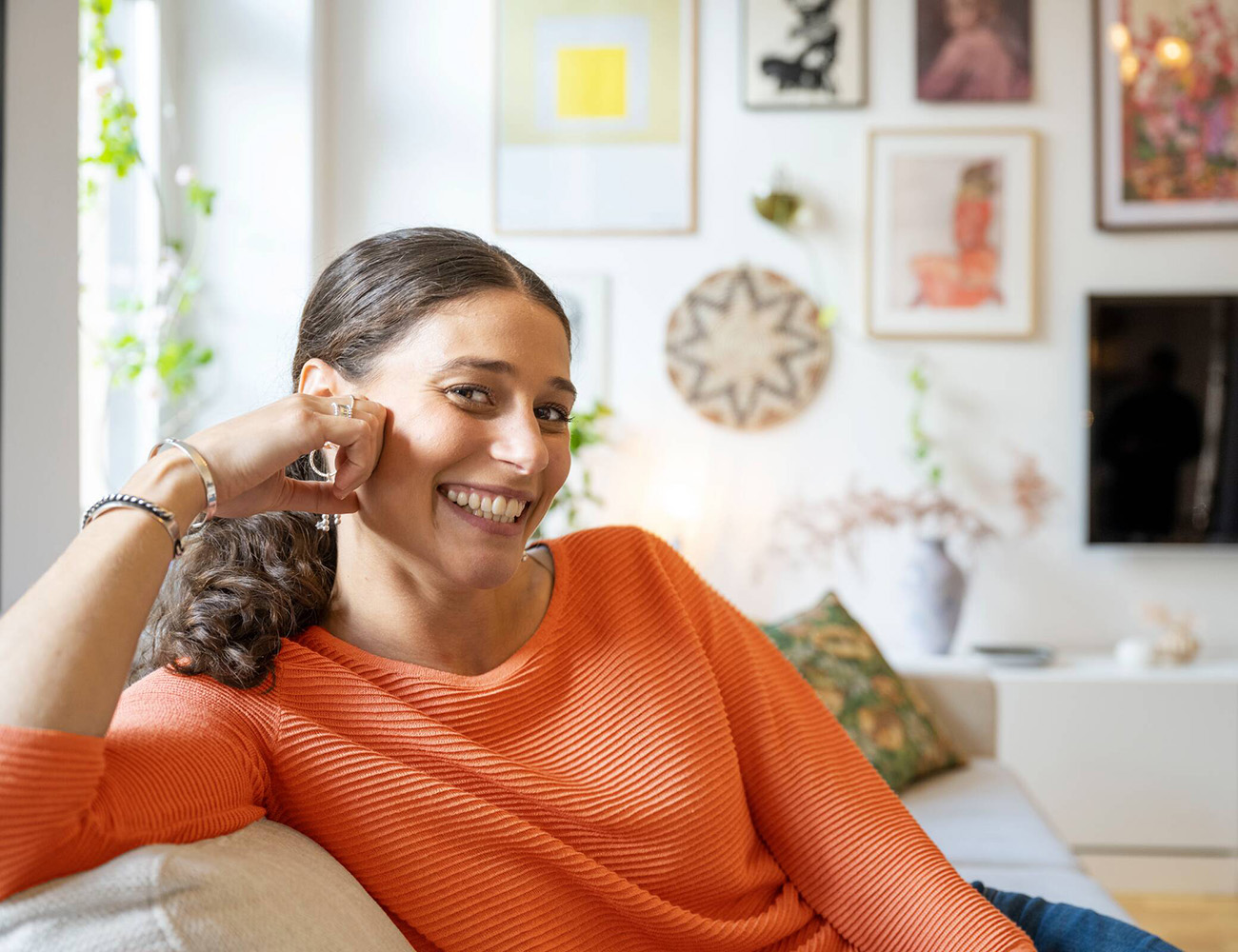 En ung mörkhårig kvinna som har håret uppsatt sitter i en vit soffa. Hon har på sig en orange tröja och sitter med armen uppe på soffstödet. Bakom henne ser man tavlor och en tv på väggen.